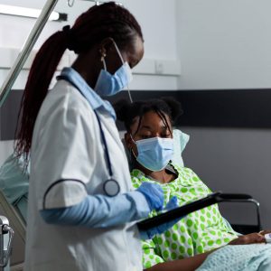 Medic of african ethnicity helping sick patient at clinic in hospital ward. Doctor using medical equipment and technology for young woman healthcare and treatment. Black person in bed